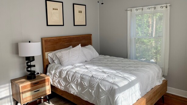 A bedroom with a wooden bed, a nightstand with a lamp, two framed pictures on the wall, and a window with a white curtain.