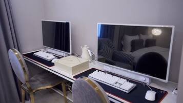 A hotel room with a desk, two monitors, a keyboard, a mouse, and a chair.