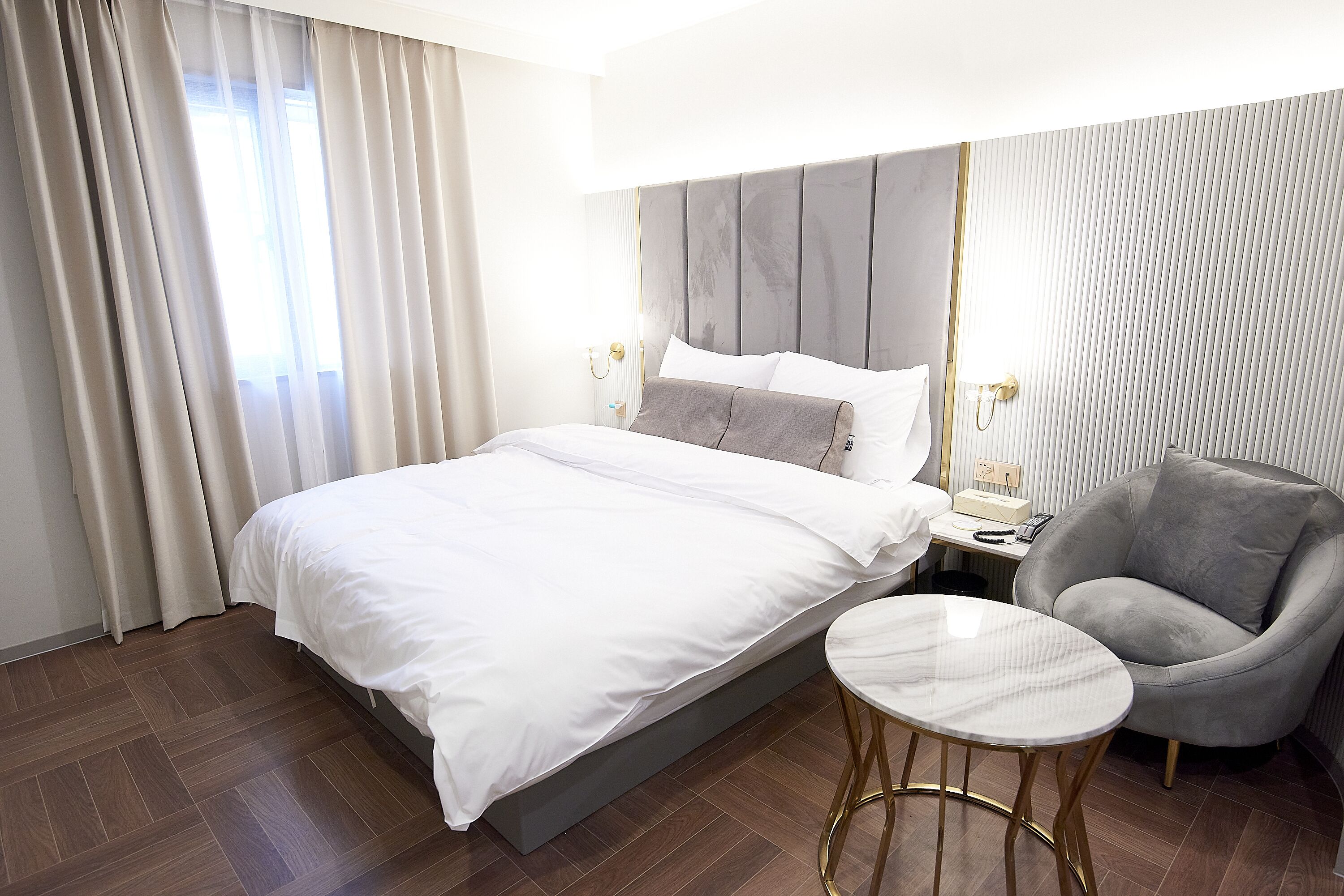 Une chambre d’hôtel avec un grand lit, un fauteuil gris, une petite table ronde et une table de chevet avec une lampe.
