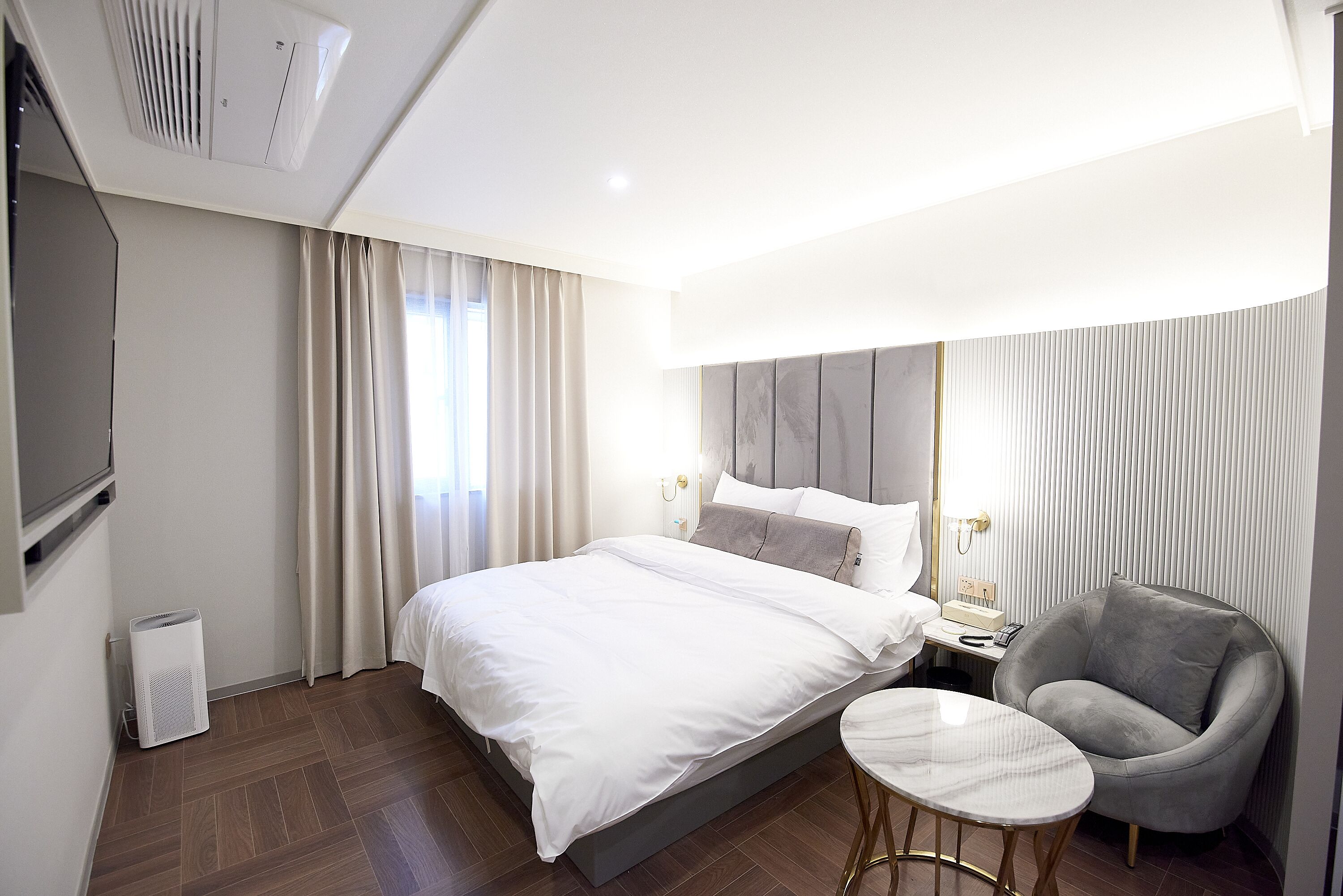 A modern hotel room with a large bed, a small round table, a grey armchair, a TV mounted on the wall, and a window with curtains.