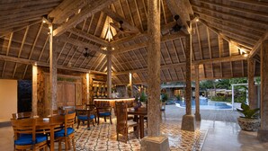 A spacious indoor area with wooden beams, a high ceiling, and a tiled floor. There are wooden tables and chairs, and a large potted plant.