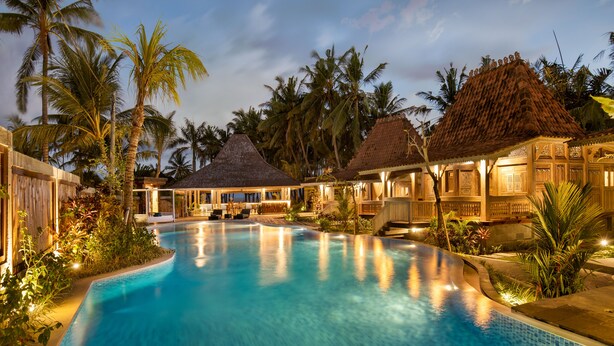 A swimming pool with a curved edge, surrounded by palm trees and thatched-roof structures.
