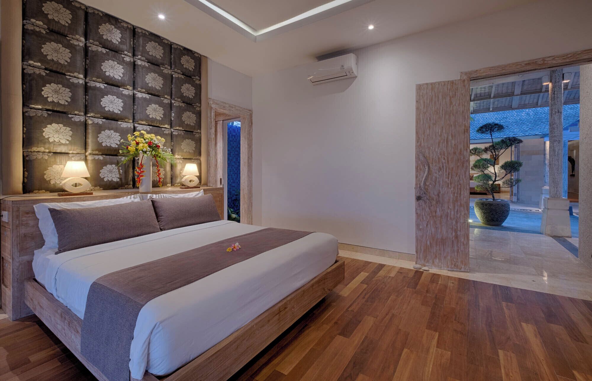 A modern bedroom with a large bed, a decorative headboard, two bedside lamps, and a view of the outdoor area.