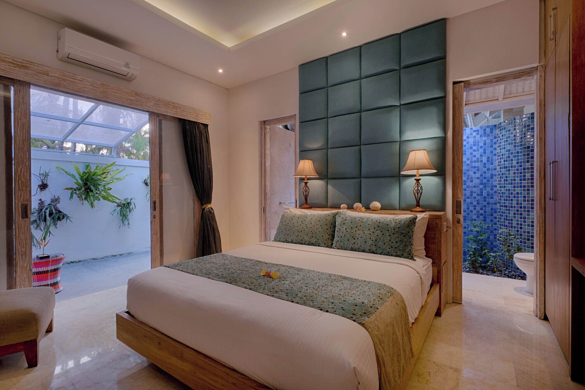 A bedroom with a large bed, two bedside lamps, a green headboard, and a view of the outside through a glass door.