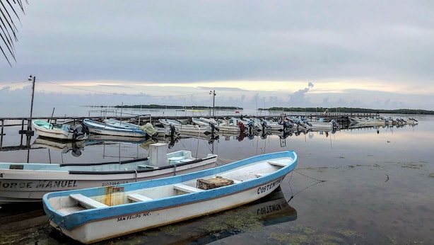 Um porto com vários barcos atracados, um cais que se estende por águas calmas e um céu nublado ao fundo.