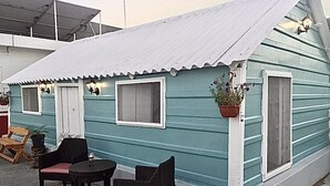 Um pequeno edifício térreo com cobertura de metal corrugado e fachada azul clara. Possui duas janelas com molduras brancas, uma porta e um pequeno alpendre com duas cadeiras de vime e uma mesa.