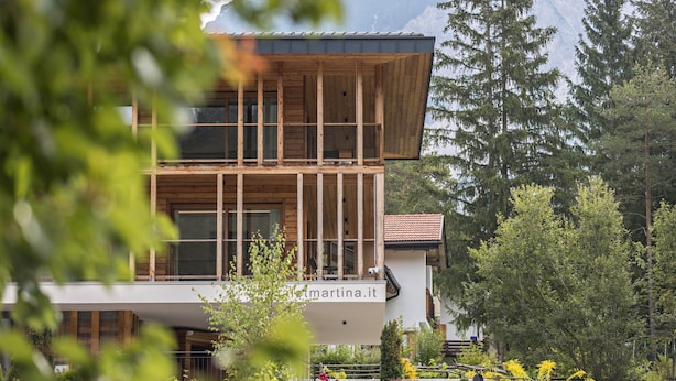 Un edificio moderno su due piani con facciata in legno e ampie finestre, immerso nel verde e circondato da montagne.