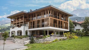 Un edificio moderno a due piani con facciata in legno e ampie finestre, circondato da un muro in pietra e vegetazione.