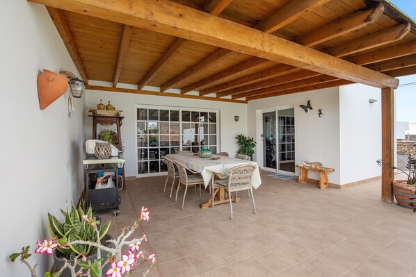 A covered patio with a wooden ceiling, a dining table with chairs, potted plants, and a window with a view.
