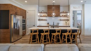A modern kitchen with wooden cabinets, a stainless steel refrigerator, a central island with bar stools, and pendant lighting.