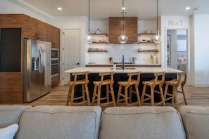 A modern kitchen with wooden cabinets, a stainless steel refrigerator, a central island with bar stools, and pendant lighting.