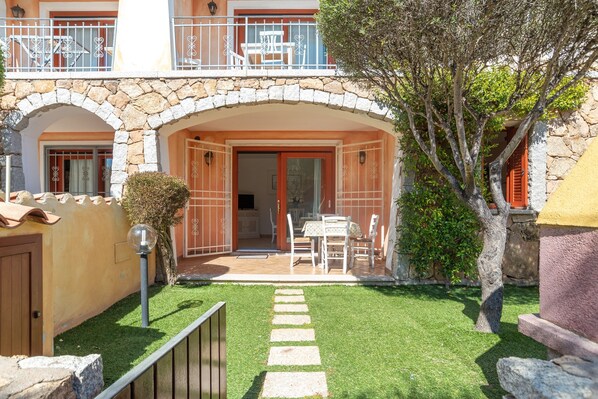 A two-story building with a stone archway entrance, a balcony with white railings, and a small patio area with a table and chairs.