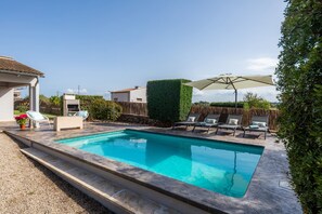 Une piscine rectangulaire bordée d’une terrasse en pierre, entourée de chaises longues et d’un parasol.