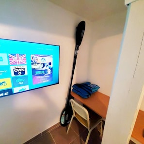A room with a flat-screen TV mounted on the wall displaying various streaming services. There is a wooden desk with a chair and a black paddleboard leaning against the wall.