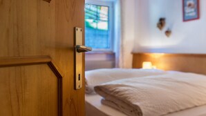 A hotel room with a wooden door, a bed with white bedding, a window with a view, and a wall-mounted light fixture. - Holiday Apartment Casa Lorenz 2 with Mountain View, Wi-Fi & Garden (Canazei)