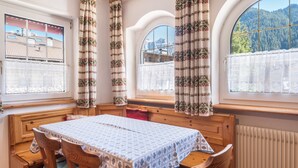 A room with a wooden table and chairs, a window with a view of mountains, and a window with a view of a building. - Holiday Apartment Casa Lorenz 2 with Mountain View, Wi-Fi & Garden (Canazei)