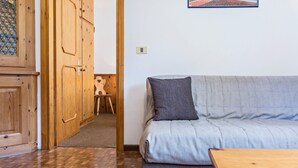 Una habitación con piso de madera, un sofá gris con un cojín, una mesa de madera y un mueble de madera con puertas de vidrio.