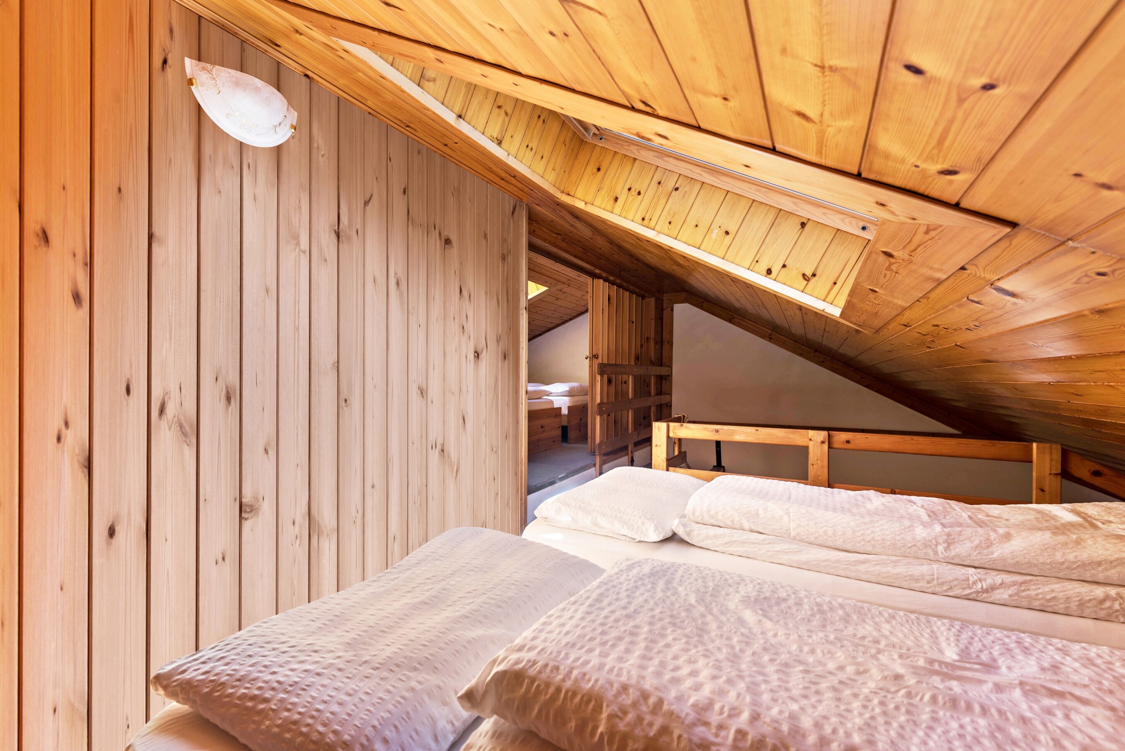 A wooden interior with a sloped ceiling, a bed with white bedding, and a visible door leading to another room.