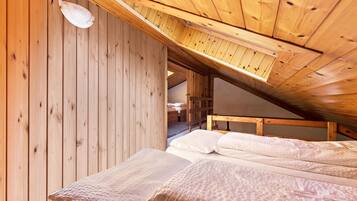 A wooden interior with a sloped ceiling, a bed with white bedding, and a visible door leading to another room.