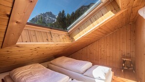 Eine Holzhütte mit geneigtem Dach, zwei Betten mit weißer Bettwäsche, ein kleiner Holzschemel und Blick auf Berge durch ein Oberlicht.