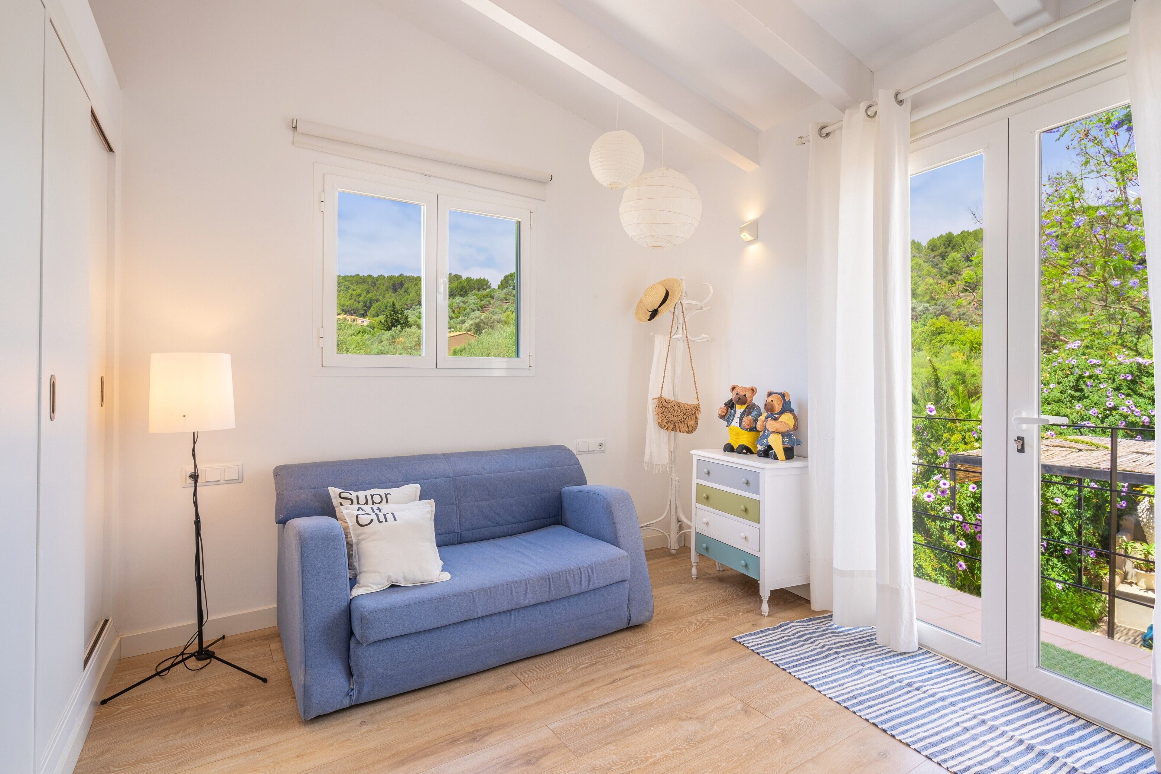 Una sala de estar con un sofá azul, una lámpara, un aparador blanco y vistas a la vegetación a través de las ventanas.