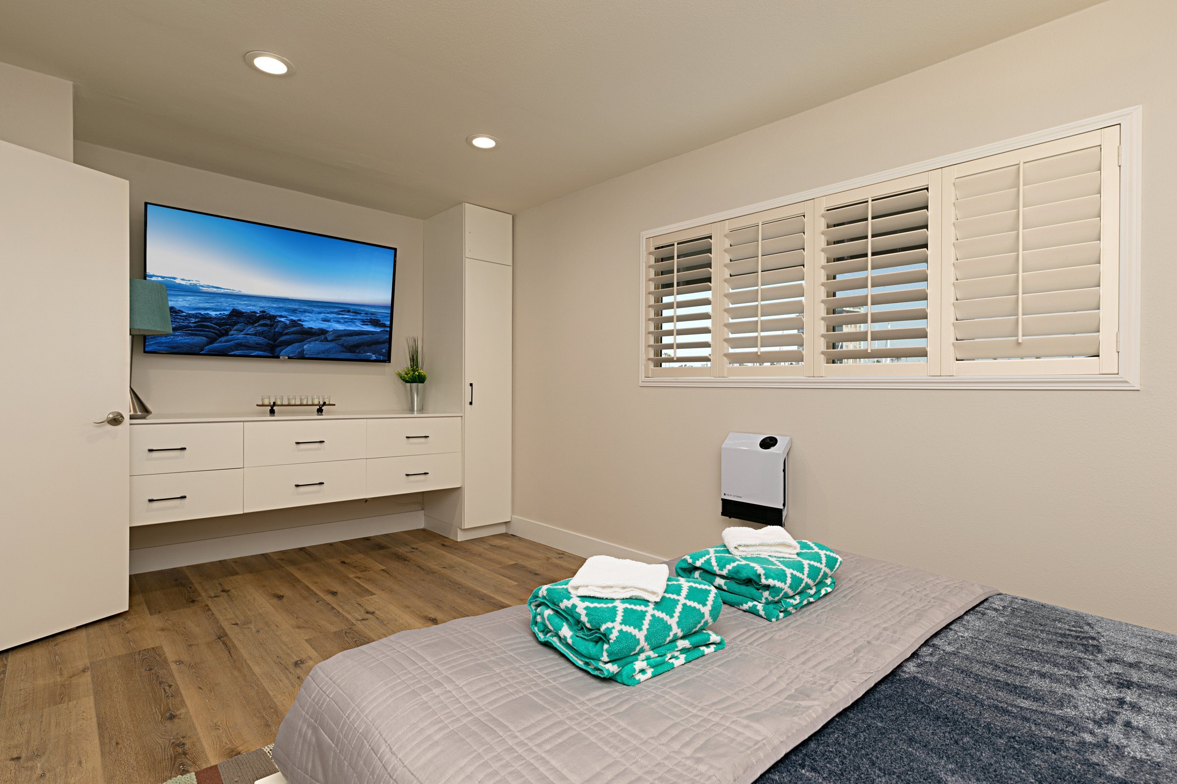 A bedroom with a bed, a TV mounted on the wall, a window with shutters, and a wooden floor.