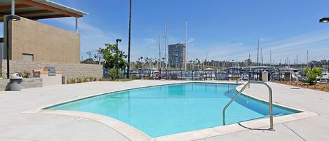 A swimming pool with a metal ladder, surrounded by a concrete deck and palm trees.