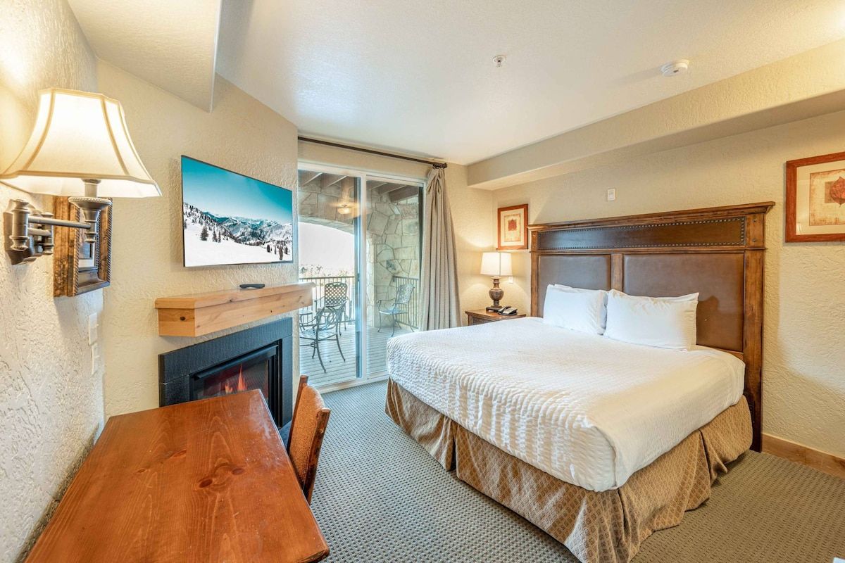 A hotel room with a bed, a fireplace, a dining table, and a view of the outdoors.