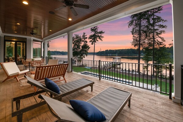 A covered outdoor area with lounge chairs and a view of a lake at sunset.