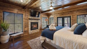 A cozy log cabin bedroom with a fireplace, a bed with pillows, a TV mounted above the fireplace, and a ceiling fan.