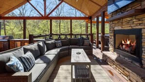 A covered outdoor patio with a stone fireplace, a large sectional sofa, and a ceiling fan.