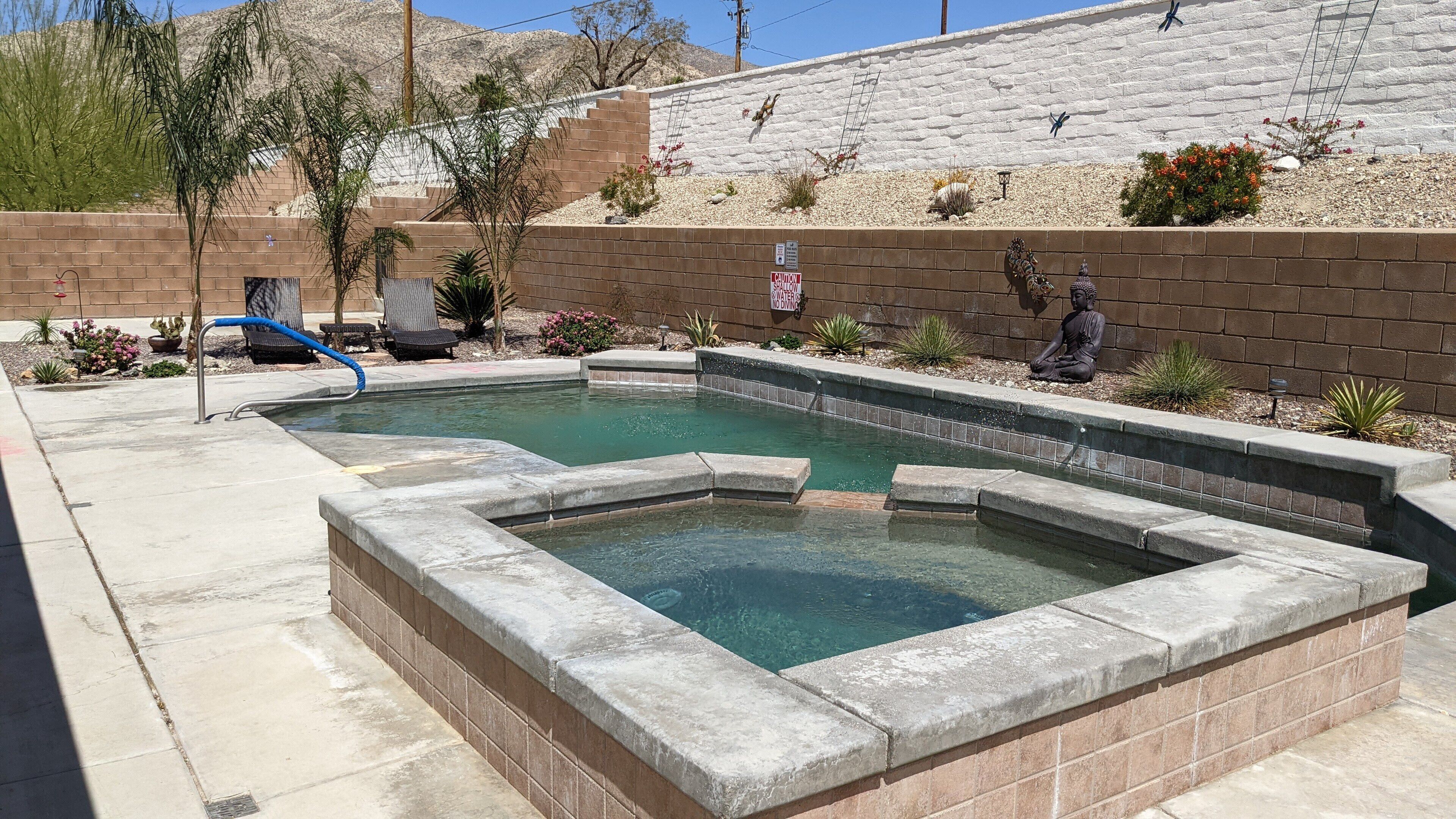 Una piscina rectangular con borde de piedra, rodeada por un muro de ladrillo y diversas plantas.