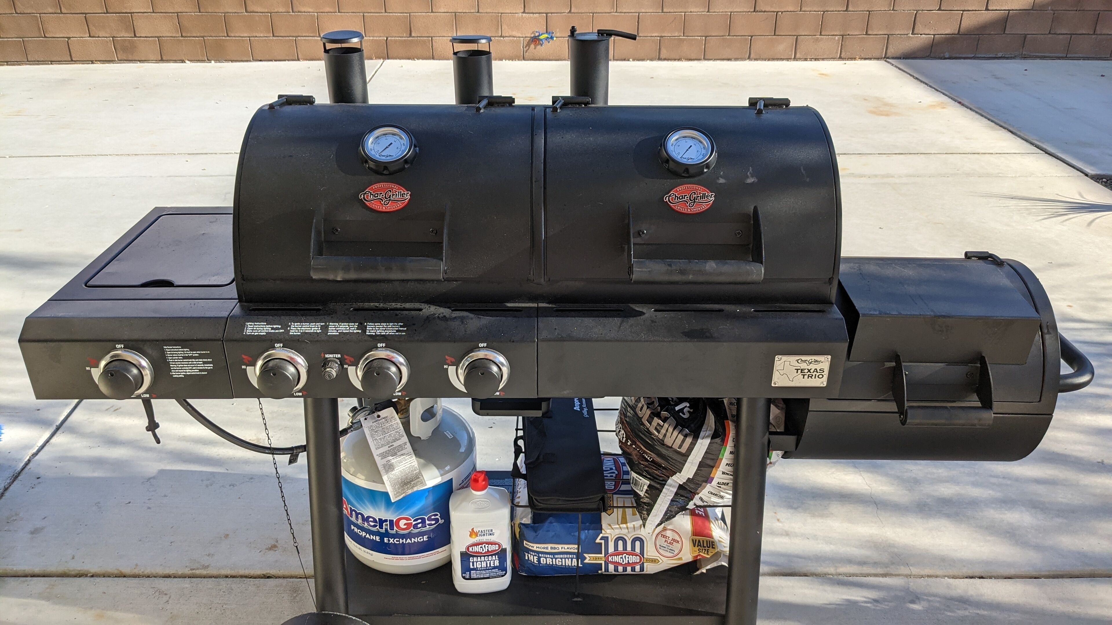 A black charcoal grill with two gauges, a propane exchange bottle, a Kingsford lighter, and a Texas Trio label.