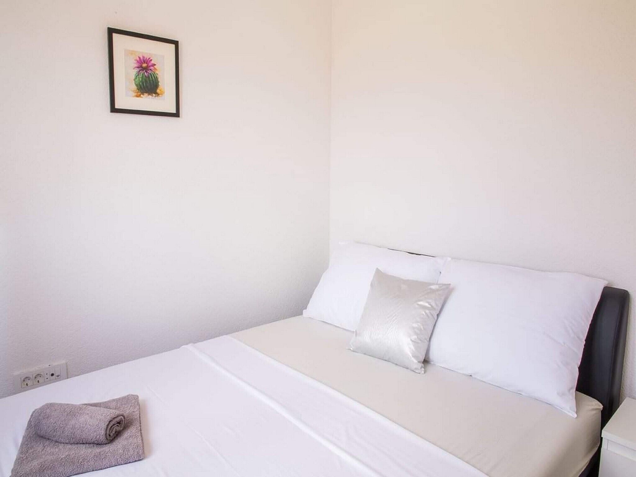 A neatly made bed with white linens and a gray towel, a framed picture on the wall, and a white nightstand with a lamp.