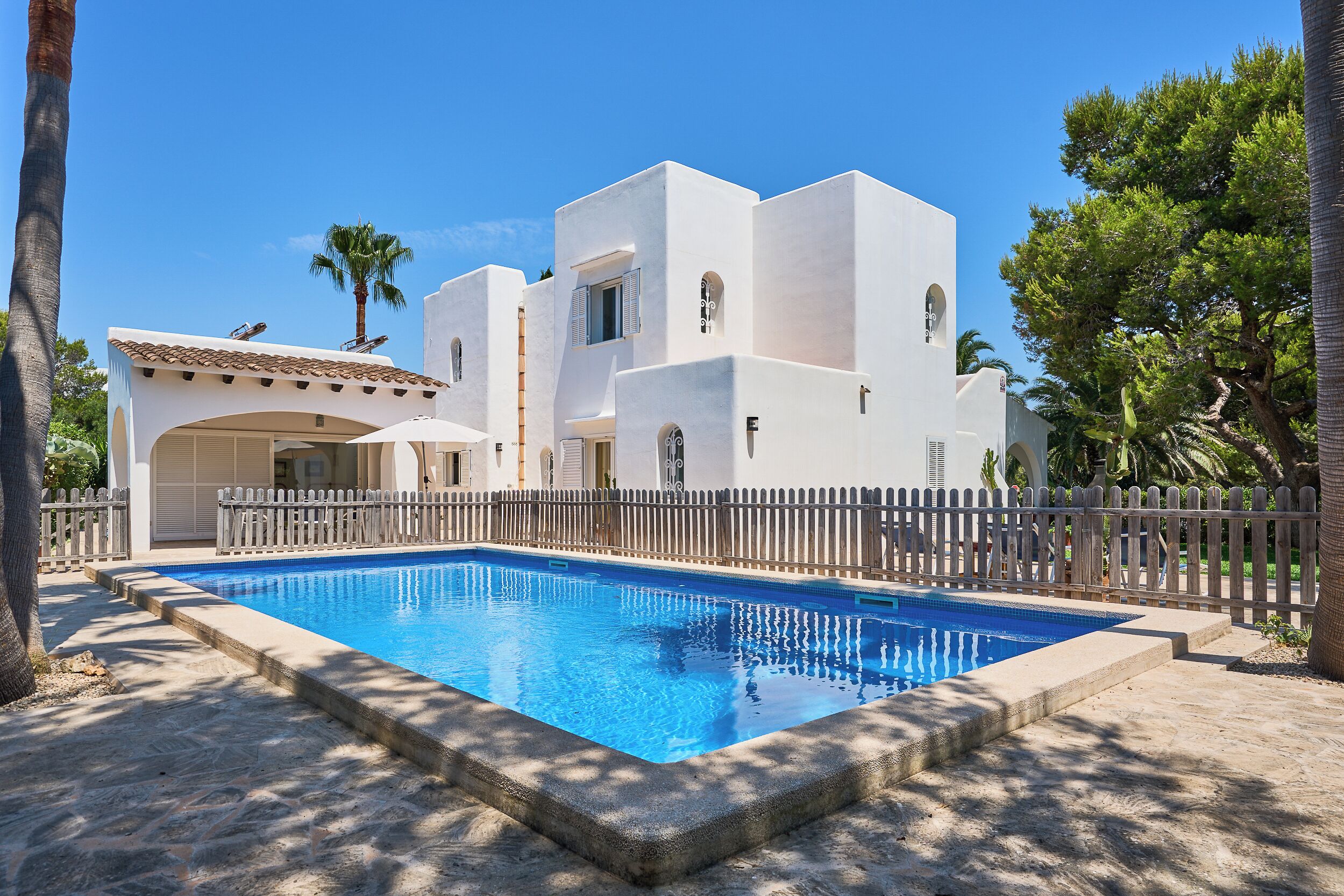 Una villa blanca con piscina, rodeada por una valla de madera y palmeras.