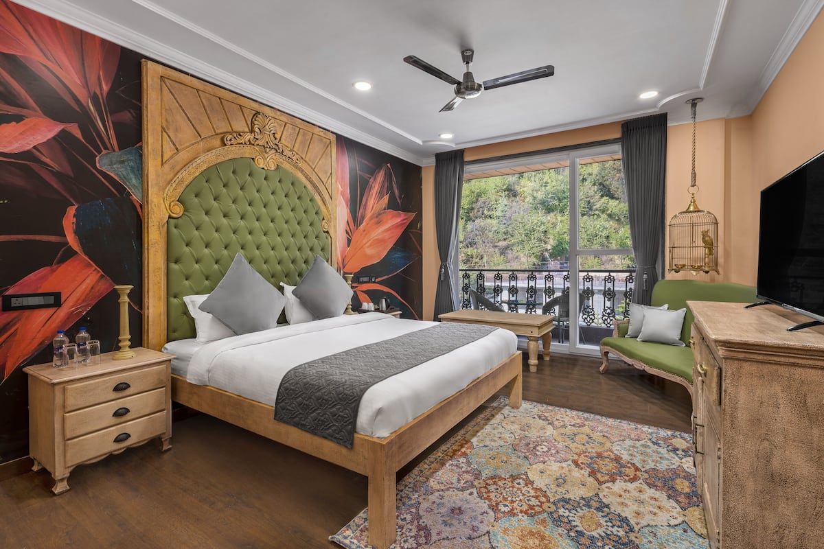 A bedroom with a large bed, a green upholstered headboard, a wooden nightstand, a green sofa, a television, and a balcony with a railing.