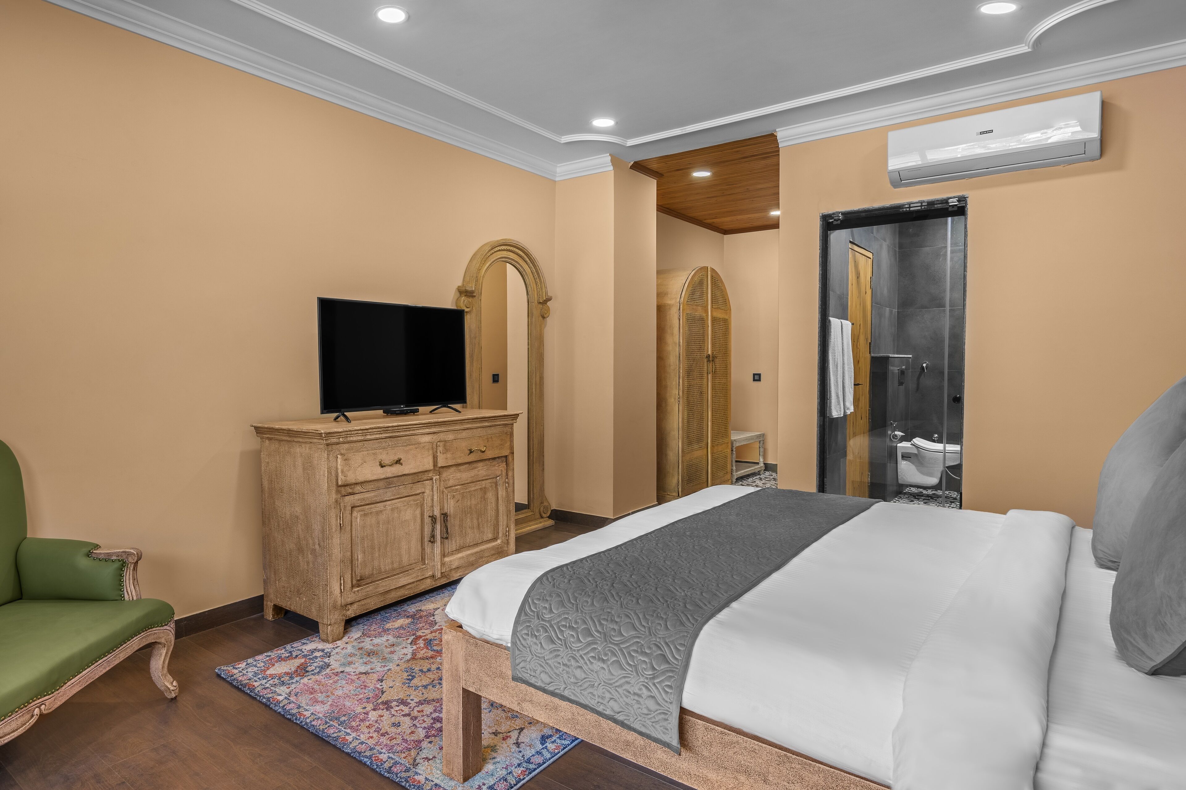 Une chambre à coucher avec un lit, un téléviseur posé sur une commode en bois, un fauteuil vert et une salle de bain visible à travers une porte en arc de cercle.