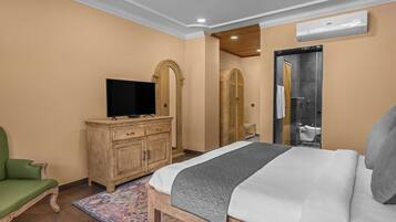 Une chambre à coucher avec un lit, un téléviseur posé sur une commode en bois, un fauteuil vert et une salle de bain visible à travers une porte en arc de cercle.