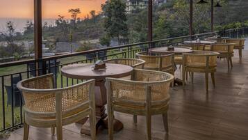 A terrace with wicker chairs and tables, overlooking a scenic landscape with hills and a body of water during sunset.