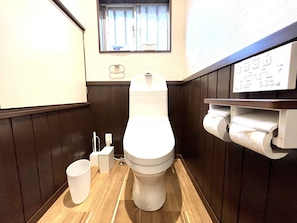 Een kleine, functionele badkamer met toilet, handdroger, papieren handdoekdispenser en raam met gordijnen.