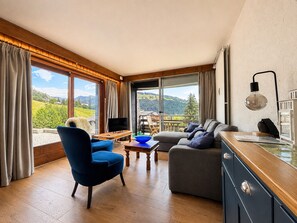 A modern living room with a large window, a blue chair, a grey sofa, and a wooden coffee table.