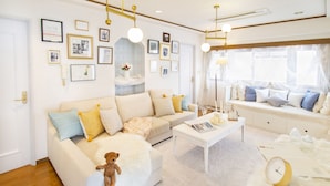A modern living room with a white sofa, a coffee table, and a teddy bear. There are framed pictures on the wall and a window with sheer curtains. - HARMONIA Tokyo Horikiri (Tokyo)