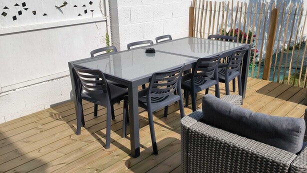 Eine Terrasse im Freien mit einem Glastisch und Stühlen, umgeben von einem Holzdeck und einer weißen Wand mit geometrischen Ausschnitten.