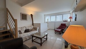 A living room with a sofa, a coffee table, a red chair, a lamp, a staircase, and a window with blinds. - Cozy 3-Level House in La Baule, 2 Terraces, Near Beach & Shops, Sleeps 6 (La Baule)