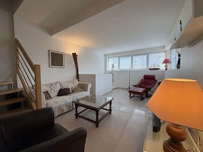 Ein Wohnzimmer mit einer Couch, einem Couchtisch, einem roten Sessel, einer Lampe, einer Treppe und einem Fenster mit Jalousien.