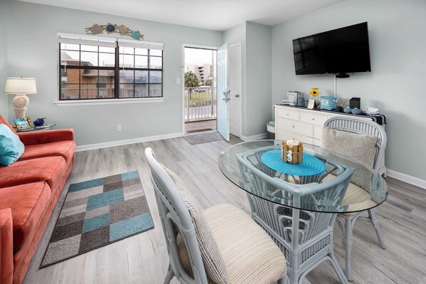 A living room with a sofa, a glass table with chairs, a TV, and a window with blinds.