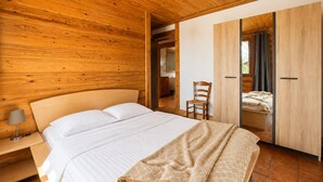 Quarto com teto de madeira, cama com roupa de cama branca, uma cadeira e um guarda-roupa.