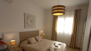 A neatly arranged bedroom with a bed, bedside lamps, a decorative wall piece, and a window with curtains. - Villa Las Tres J (Torrox Costa)