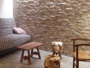 Un salon avec un mur en pierre, un canapé à motifs, une table basse en bois, un petit tabouret et une chaise en bois.