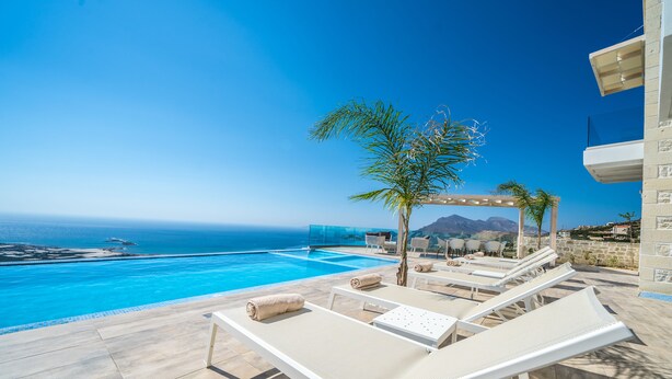 Ein Poolbereich mit Liegestühlen, einer Palme und Blick auf das Meer und die Berge.